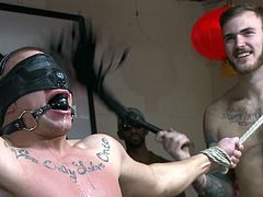 The gay slave hunk is tied to a pole in the sex club and the gay dominators, are making him endure all sorts of torture. They attach clamps to his nipples and this makes him scream, but his screams are muffled, because of is gag. He gets clamps on his scrotum and that hurts even more.